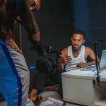 Dois jovens gravam um vídeo em um estúdio doméstico, com câmera, laptop e outros equipamentos de produção ao redor.