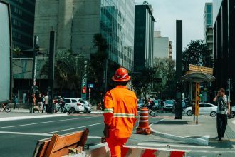 Trabalhador da construção civil em uma movimentada rua de São Paulo, cercado por edifícios e circulação urbana.