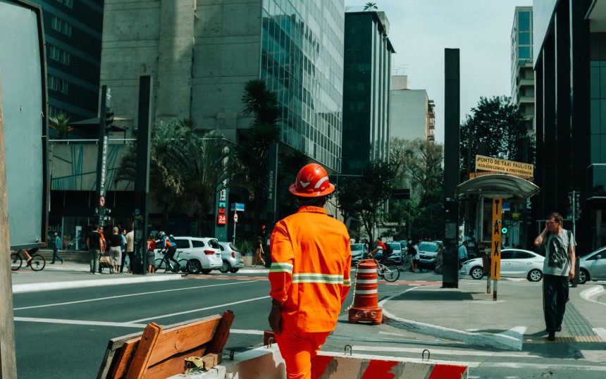 Trabalhador da construção civil em uma movimentada rua de São Paulo, cercado por edifícios e circulação urbana.